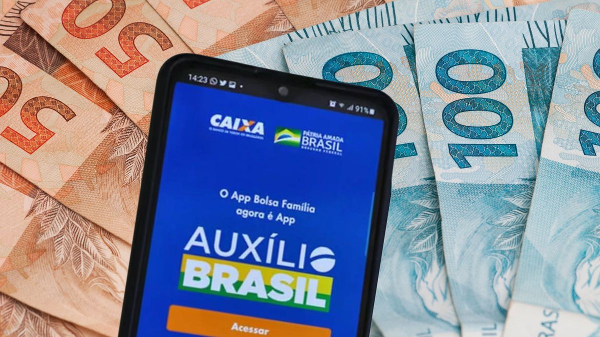 Um celular aberto com o programa do Auxílio Brasil ao lado de notas de dinheiro