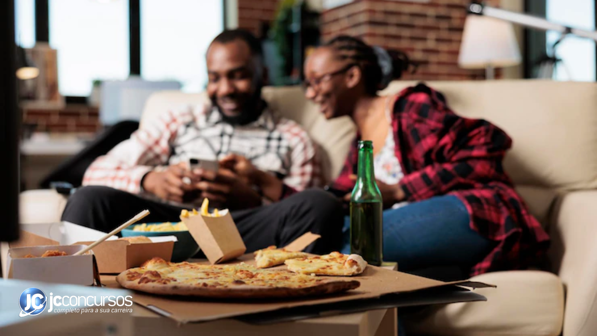 Casal sorrindo olha para o celular, sentados em frente à uma mesa com pizza e garrafa de vidro verde