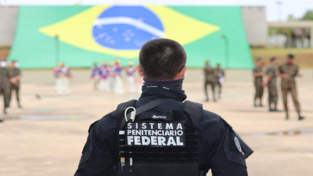 Concurso Depen: com bandeira do Brasil ao fundo, agente de segurança aparece de costas observando militares