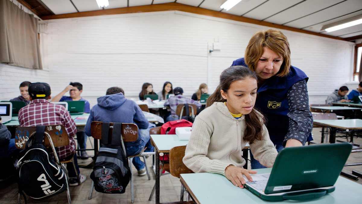 Concurso Prefeitura de Candelária: estudante utilizando computador recebe instruções de professora