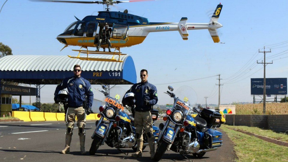 Concurso PRF: com helicóptero ao fundo, dois agentes da corporação posam para foto ao lado de motos