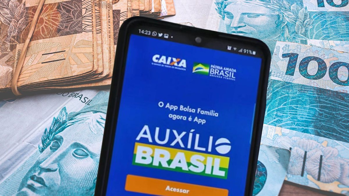 Um celular aberto com o programa do Auxílio Brasil ao lado de notas de dinheiro