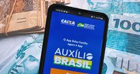 Um celular aberto com o programa do Auxílio Brasil ao lado de notas de dinheiro - Divulgação - Empréstimo Auxílio Brasil
