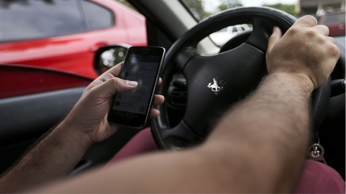 Um motorista com a mão no volante enquanto segura um celular