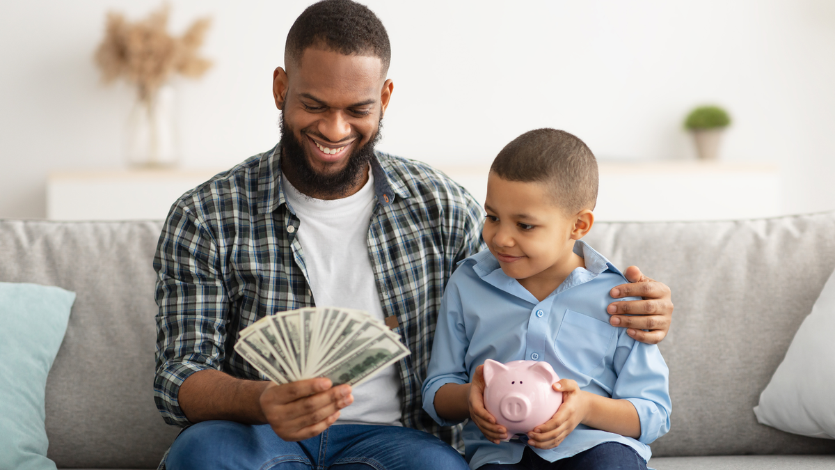 Pai abraça filho e segura notas de dinheiro enquanto criança segura cofrinho em formato de porco