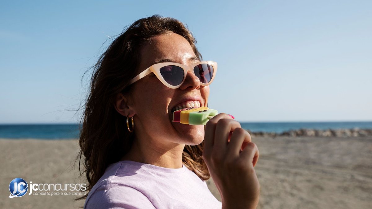 Mulher tomando picolé na praia enquanto usa óculos escuros